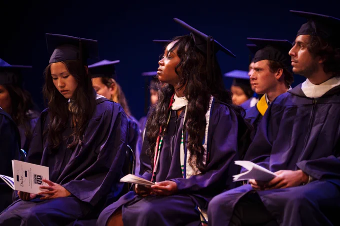 Seated students in deep blue undergraduate caps and gowns look at their programs and at the speaker during the English Department's diploma ceremony