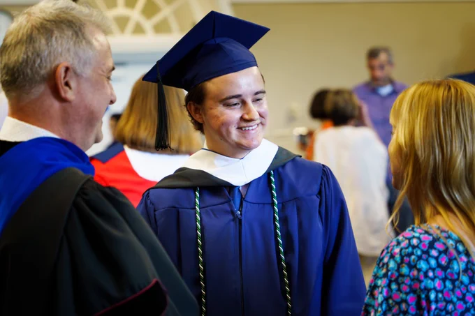 A graduating Philosophy student in a dark blue cap and gown with a cord smiles during a conversation with a diploma ceremony guest and a professor