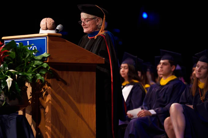A brain sits atop a wooden lectern as a faculty member speaks in her academic regalia, with graduating students seated behind her