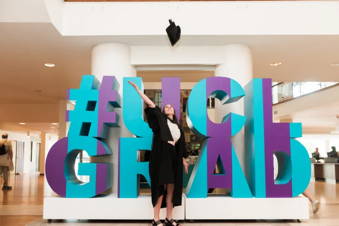 A graduate throwing her hat up in front of large letters saying #UCLGrad
