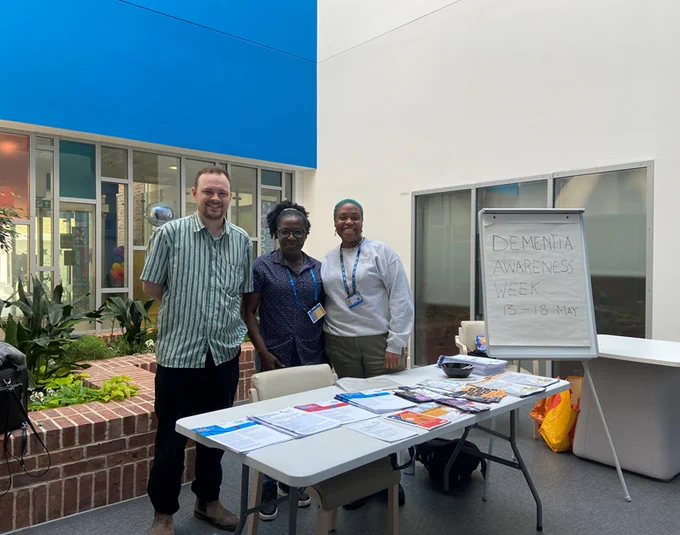 Three people standing behind a table covered with information leaflets and flyers. To the side of them is a flipchart easel with a handwritten sign which reads: Dementia Awareness Week, 13-18 May.
