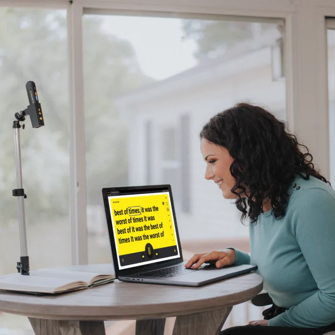 A woman using OrCam's Smart Magnifier on her computer screen, with the OrCam Read 3 positioned in its stationary stand beside her, aimed at a book.
