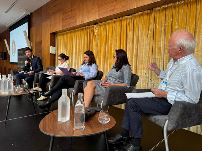 Image of 5 speakers sat on the stage. Two male speakers sit on either end and three female speakers sit in the middle.