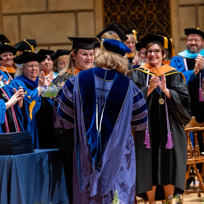 Kaleigh Sullivan acceptes a bouquet of flowers from Dean Lisa Kitko during graduation. Professors applaud her in the background.