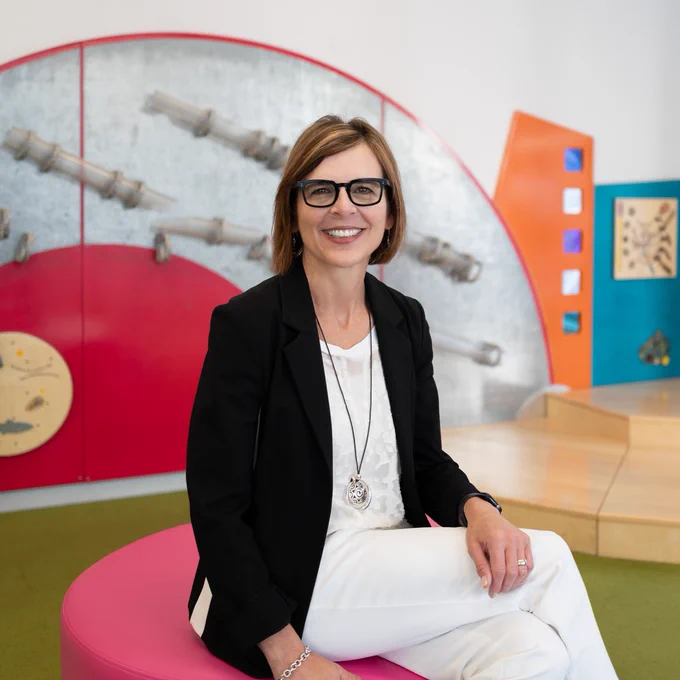 ID 1: Photo of a person with short blonde hair, wearing black glasses, a black blazer, and white pants, sits on a pink stool with their legs crossed. Behind them is a colorful, playful library space. 