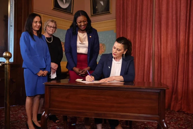 Governor Gretchen Whitmer sits at a table signing a piece of legislation whole Anita Fox, Marianne Huffs, and Sarah Anthony look on.