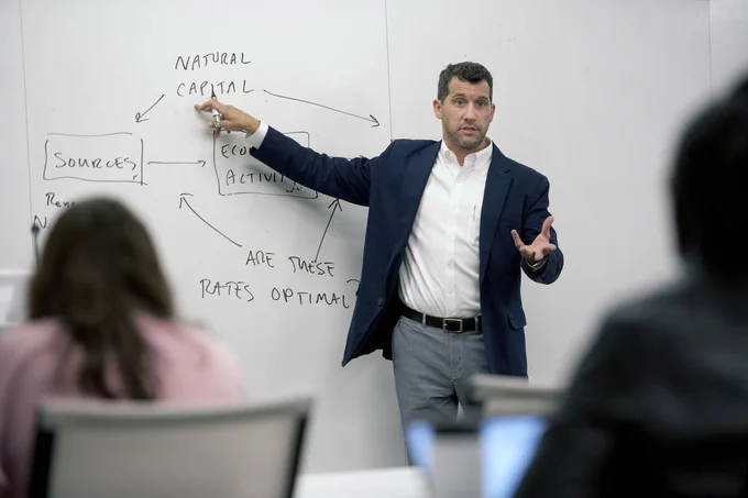 Man standing at the front of a class with a drawing on a white board that includeds the words "natural capital," "sources," "are these rates optimal" connected by arrows.

