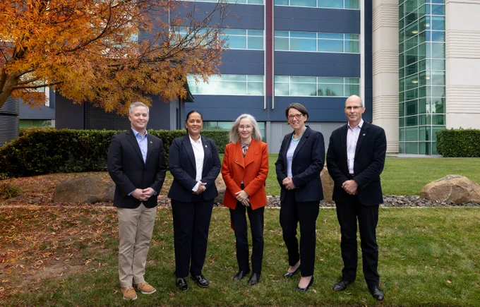 Five people in business attire stand in front of a building with an orange-leafed tree in between. The five people are a man in a navy suit, a woman in a black suit, a woman in a red jacket, a woman in a navy suit, and a man in a black suit.