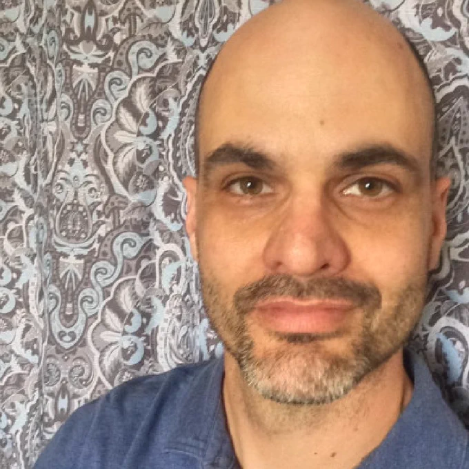 Dr. Deman with a shaved head and stubble smiling slightly at the camera, wearing a blue shirt, with a patterned background.