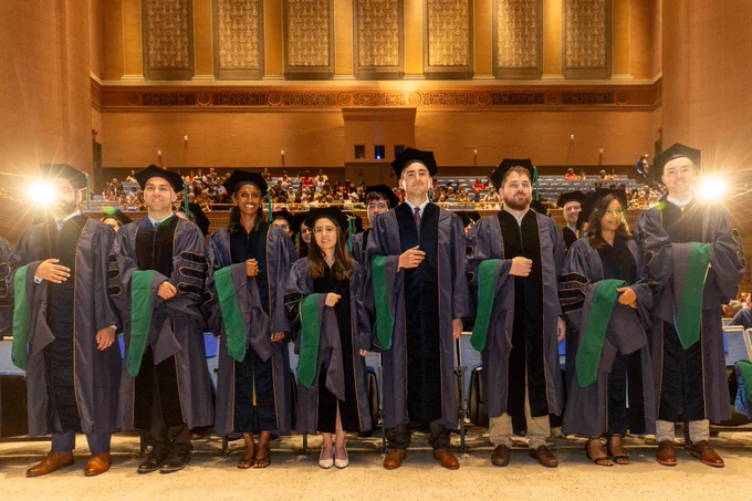 School of Medicine Class of 2024 graduates in regalia standing and smiling in the Soldiers and Sailors Memorial Hall auditorium.  