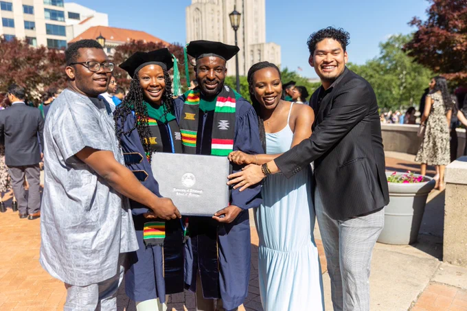 Two School of Medicine Class of 2024 graduates in regalia smiling for a photo in front of the Cathedral of Learning with three friends. 