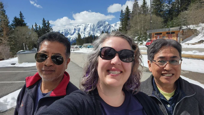 Three happy people smiling into the camera with snowy mountain peaks and pine forest in the background. 