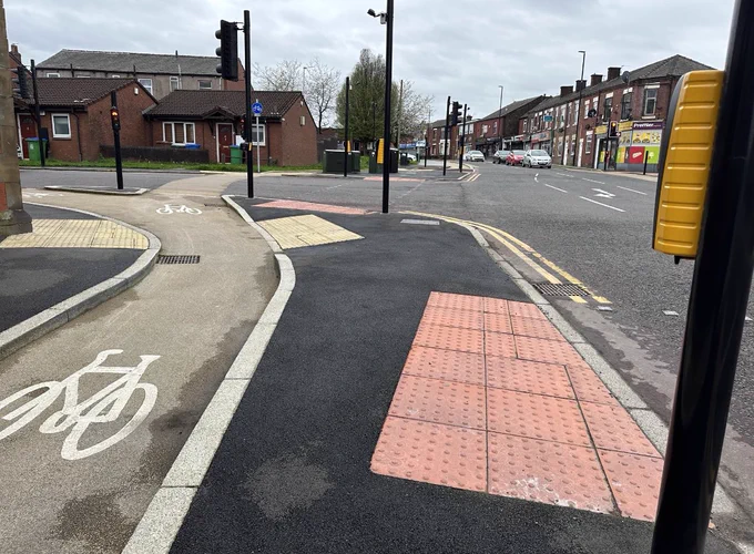 The pavement has a cycle lane running through the middle which is has kerbs either side. There is a dropped kerb with beige tactile on the corner of the pavement which crosses over the cycle lane (beige colour) onto a tarmaced island. The elongated island has a controlled crossing on one end and another one on another side of the junction. Both have red tactile laid where it meets the road. However one of the controlled crossing points there is no tactile where it is level surface with the road which is very dangerous. On this controlled crossing there is just one pole with a push button crossing on it.