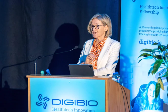 A woman wearing glasses and a white blazer with an orange patterned dress is speaking at a podium. The podium has the "DIGIBIO" logo and text about the Healthtech Innovation Fellowship. There is a banner in the background with additional information about the program.