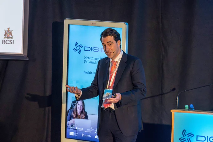 A man in a suit and pink tie is speaking on a stage. He is gesturing with his hands, and there is a screen behind him displaying the "DIGIBIO" logo and information about the Healthtech Innovation Fellowship.