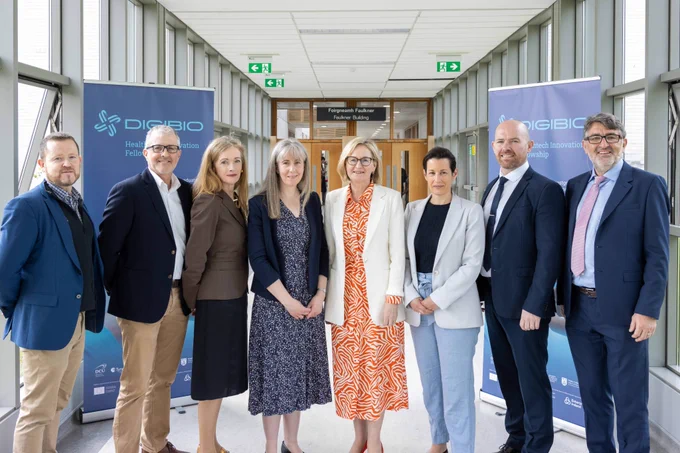 A group of eight people stand in a hallway with large windows. They are dressed in professional attire and smiling at the camera. Behind them, there are two banners displaying the "DIGIBIO" logo and text related to the "Healthtech Innovation Fellowship."
