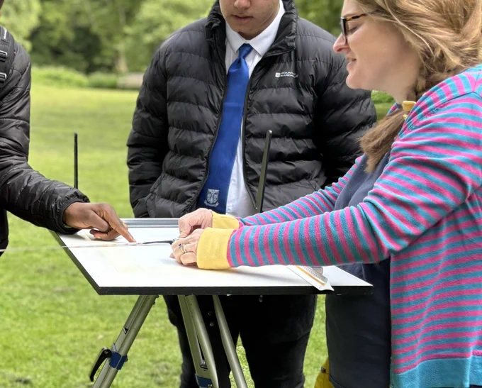 Photo showing 3 people standing around a plane table. Two students in black jackets and a female member of staff wearing a pink and turquoise striped cardigan. A tape is stretched out across the plane table board. One student is pointing to a point on the board