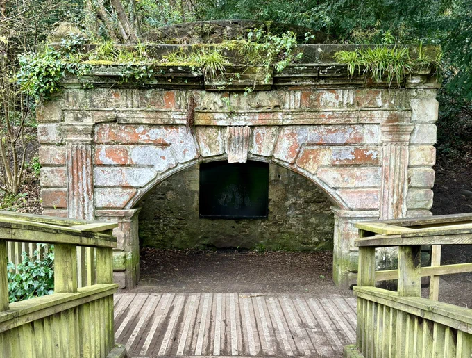 A photo showing the view down from the bridge towards the ornate stone archway to old icehouse. The metal plate covering the entrance to the icehouse can be seen behind the arch.