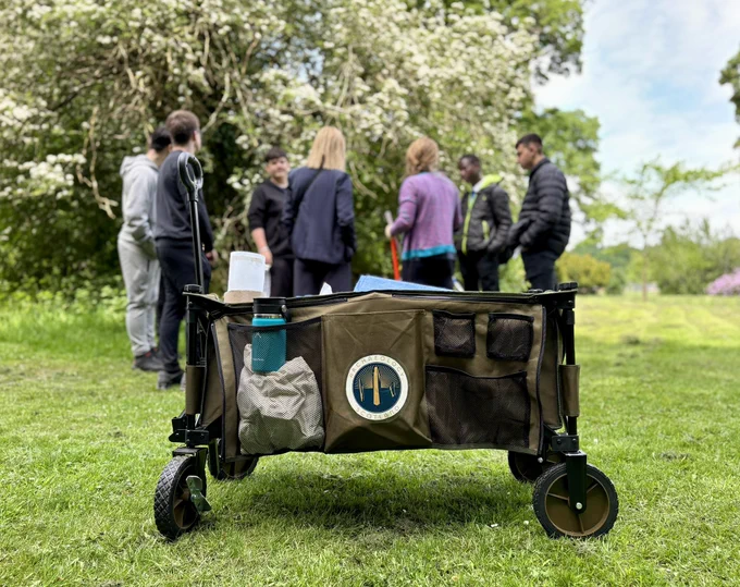 Photo showing the side view of a khaki coloured outdoor trolley with wheels on grass - Archaeology Scotland logo on the side and a turquoise water bottle in the side pocket. Behind the trolley are a group of young people standing around a plane table. Trees and bushes can be seen in the distance.