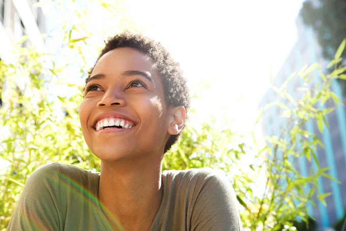 Person laughing outdoors in sunshine with blurred greenery and buildings in background.