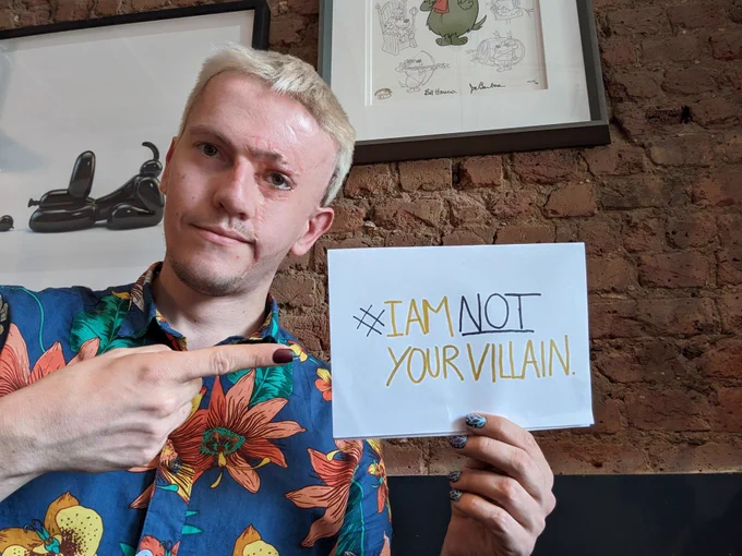 Our ambassador Rob wearing a floral shirt and pointing to a sign he's holding which reads: "#IAmNotYourVillain"
