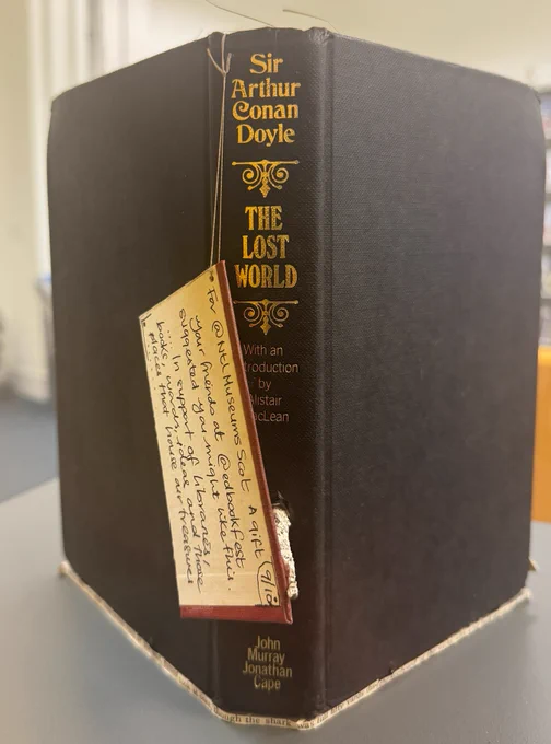 Outer cover of the book sculpture featuring dark cloth covers from a copy of  The Lost World by Sir Arthur Conan Doyle and a note that reads For @NtlMuseumsScot - a gift your friends at  @edbookfest suggested you might like this...In support of libraries, books, words, ideas and those places that house our treasures