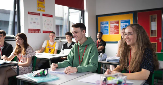 Students sitting at desks in a brightly lit classroom, smiling and looking towards a teacher out of view. The room features colorful educational posters and a whiteboard. Everyone appears engaged and attentive.