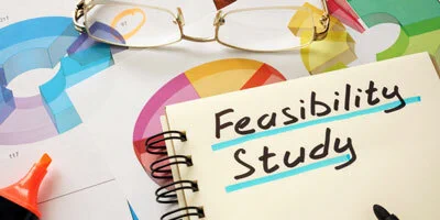 An open notebook with the words "Feasibility Study" written in bold, surrounded by colorful charts, a highlighter, and a pair of reading glasses resting on top of the page.
