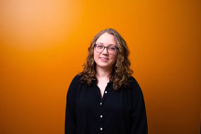 Librarian Katie Harding with shoulder-length curly brown hair wearing glasses and a black button-up shirt stands smiling in front of an orange wall. 