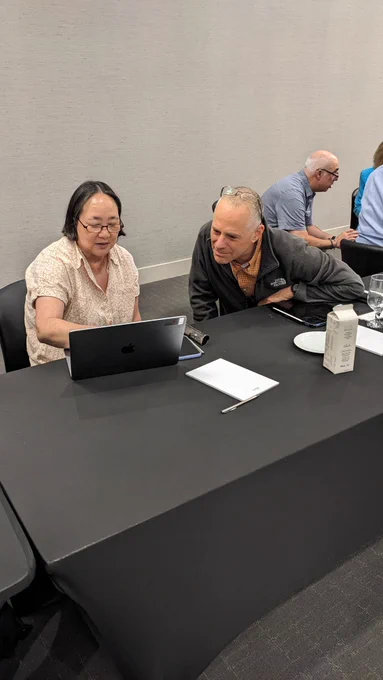 Ann West and Steve Caplan sit next to each other at a table with a black tablecloth while looking at a laptop.