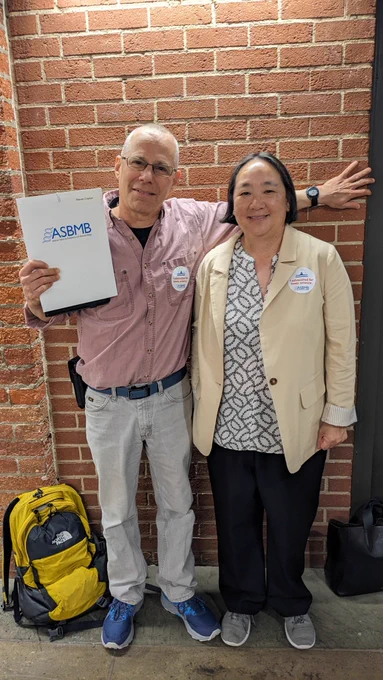 Steve Kaplan, holding a white folder, and Ann West pose for a photo in front of a brick wall in the basement of the U.S. Capitol.