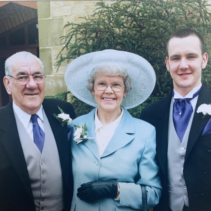 Three individuals posing for a photo outside a building. On the left, an older man wearing a dark suit and a boutonniere. In the center, an older woman in a light blue suit, black gloves, and a wide-brimmed hat, also wearing a boutonniere. On the right, a younger man in a dark suit with a boutonniere. All are smiling and looking at the camera. There is a backdrop of a stone building partially covered in ivy.