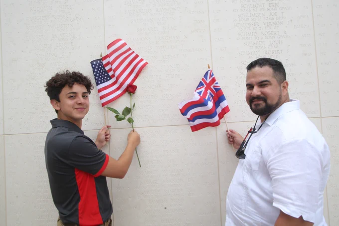 Color photograph of a young Latino teen & an adult Latino man standing in front of a stone wall with names on it. The young man on the left holds a small USA flag and a red rose. The man on the left holds a small Hawaiian flag.