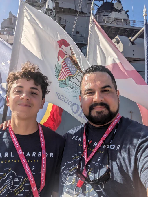 Color photograph of a young Latino man on the left and an adult Latino man on the right. They are both wearing black tshirts & red lanyards. Behind them is an Illinois flag and part of a gray battleship. 