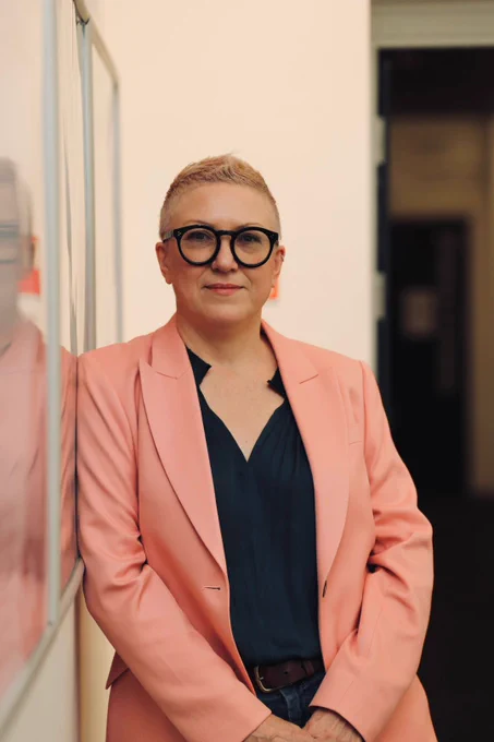 Executive Producer Rebekah Wray-Rogers leans against a white wall and looks towards the camera