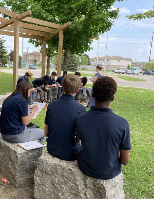 Students sitting in a semi-circle, writing down their ideas while sitting on rocks in our outdoor classroom, listening to a story read by our Learning Commons Specialist
