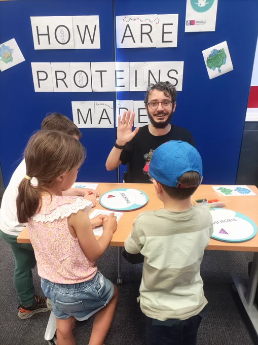 A group of children work on an activity at a table. Facing them sits a scientist, waving, in front of a board signed 'How are proteins made?'.