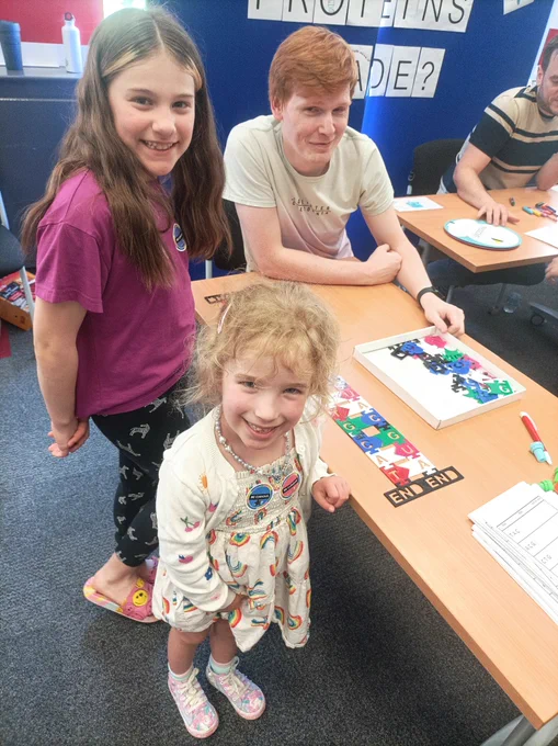 Two children stand smiling beside strings of plastic DNA and RNA bases on a table top.