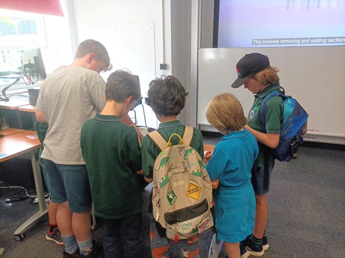 A group of children, facing away from the camera, stand around a table as they work on an activity together. 