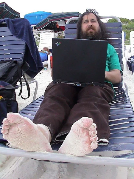 richard stallman sitting on a sunbed with his ancient thinkpad. his naked, sand-covered feet are in the foreground.
