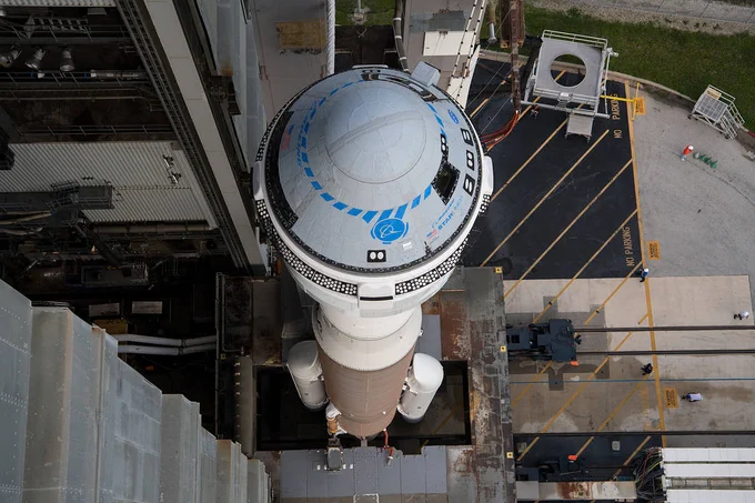 A United Launch Alliance Atlas V rocket with Boeing’s CST-100 Starliner spacecraft aboard is seen as it is rolled out of the Vertical Integration Facility. The spacecraft is silver and blue, the rocket is tan and white, and the background is gray, black, and beige. The image is shot from above the rocket looking down. Credit: NASA/Aubrey Gemignani
