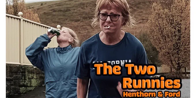 Outdoors in cold, wet weather. The trees I’m the background are brown. Two ladies are jogging. One, in the forefront has a blue t shirt on, dark, thick rimmed glasses and a cigarette in her mouth. She has short ginger-blonde hair. She looks in pain. The Second Lady just behind the first has a blue waterproof jacket on. She has her head back and is swigging from a can of Gin and Tonic.