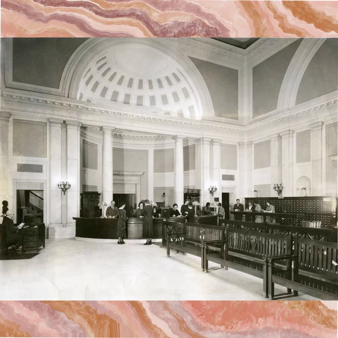 A black and white photograph of the interior of a large marble building. There are desks, including a larger central desk, with people in period dress congregating around. There are rows of wooden benches on the right-hand side of the room and a large card catalog behind them. The background of the post is a Marbleized Geode Pink and Brown Wavy Stone background.