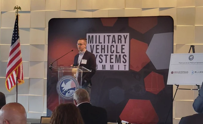 A man speaks at a podium in front of a sign that reads "Military Vehicle Systems Summit." An American flag is on the left, and audience members are seated in the foreground.