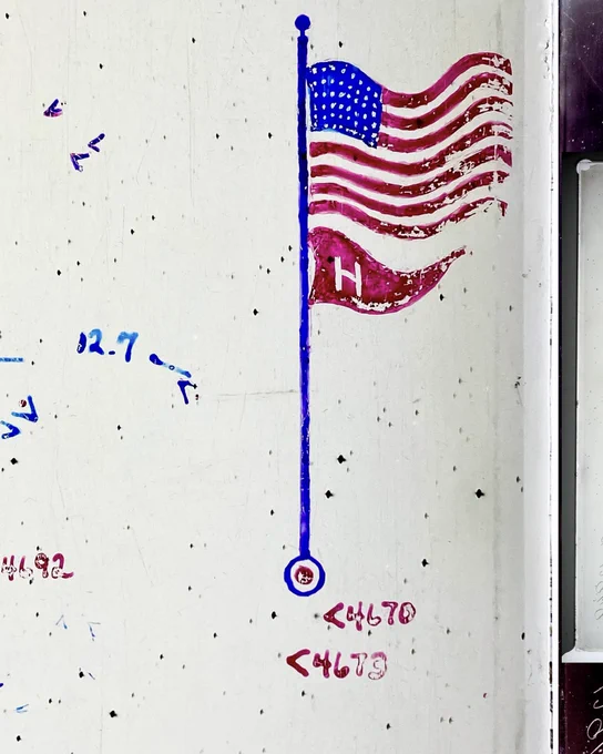 A close-up view of a flaking ink drawing of an American flag with a smaller red Harvard "H" pennant flag on a blue flag pole. This is on the edge of a black-and-white negative of the night sky, with other annotations and numbers pointing at black dots of stars or galaxies.
