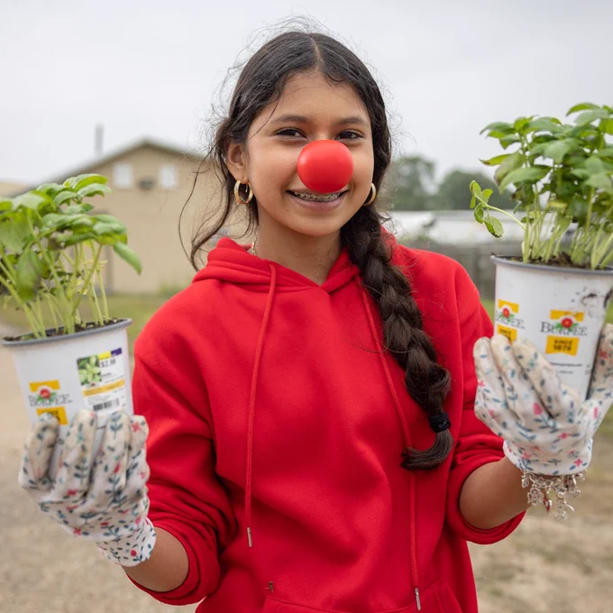 A young girl holds up a pot in each hand, which both have small, leafy green plants in them. Her long, dark hair is in a braid, and she is wearing a red sweatshirt and red nose for "Red Nose Day".