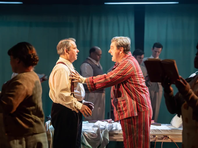 A man wearing striped pyjamas holds onto another man. Behind them there are hospital beds and people read books.