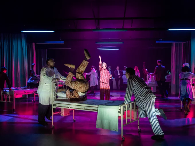 Michael Sheen wearing striped pyjamas stands in the middle of a room surrounded by hospital beds. Patients dressed in pyjama, nurses and doctors push these beds around the room.