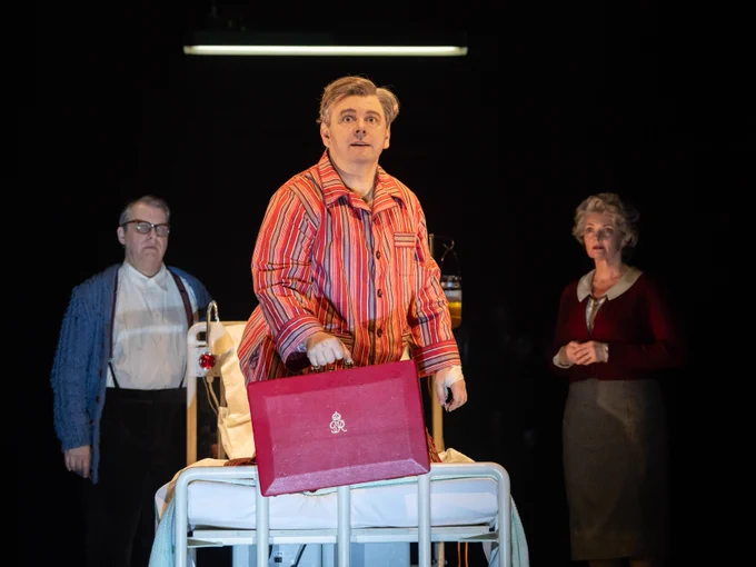Michael Sheen holds a red briefcase and wears striped pyjamas as he stands on a hospital bed. Behind him stand a man and a woman.