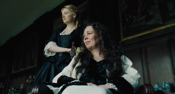A film still from The Favourite. A woman with long dark hair sits in a wooden wheelchair. She stares ahead with a small smile on her face, her eyebrows slightly raised. The woman behind her has blonde hair, wears a dark dress, and has a hand on the wheelchair. She holds a similar expression as she stares ahead.